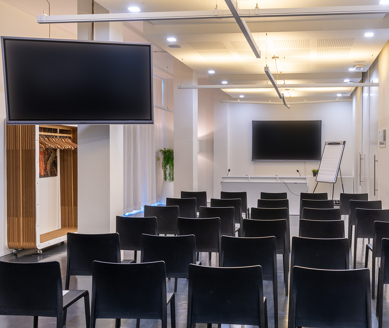 Sala conferenze con sedie nere, grande schermo, schermo aggiuntivo laterale e lavagna a fogli mobili.