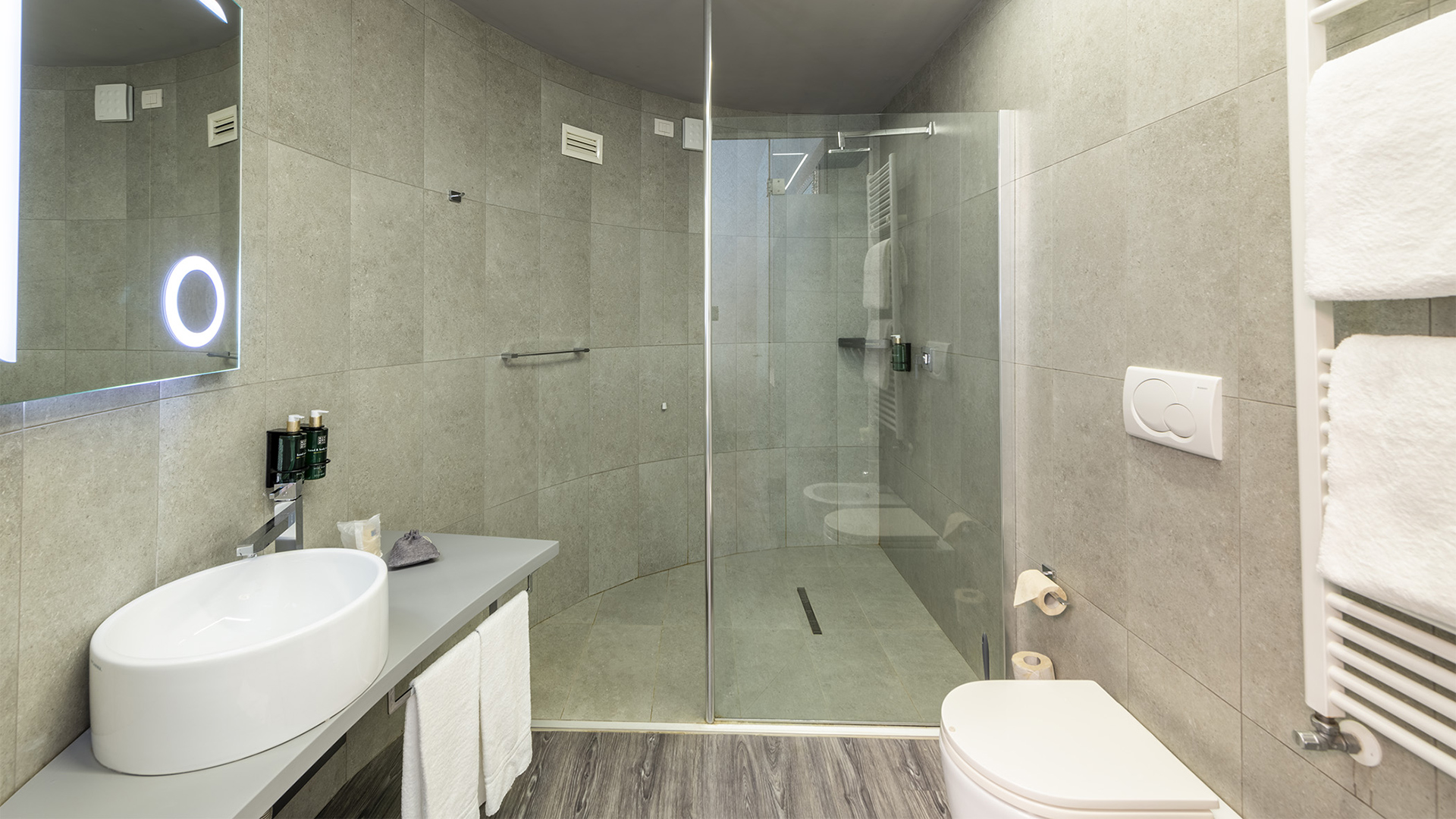 Bagno Family Room con ampio box doccia in vetro, lavabo da appoggio su piano grigio, specchio retroilluminato, sanitari sospesi e rivestimenti in gres effetto pietra