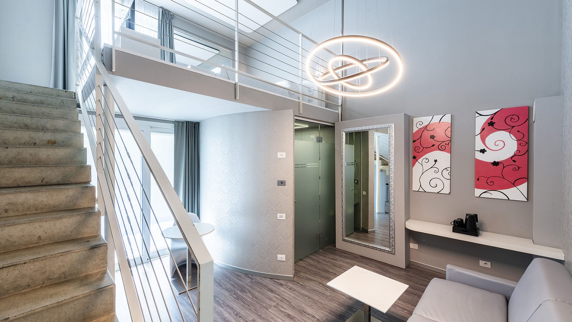 Junior Suite Deluxe su due livelli con scala interna, soppalco notte con ringhiera bianca, zona living con divano e tavolino, specchio a parete e lampada a sospensione di design