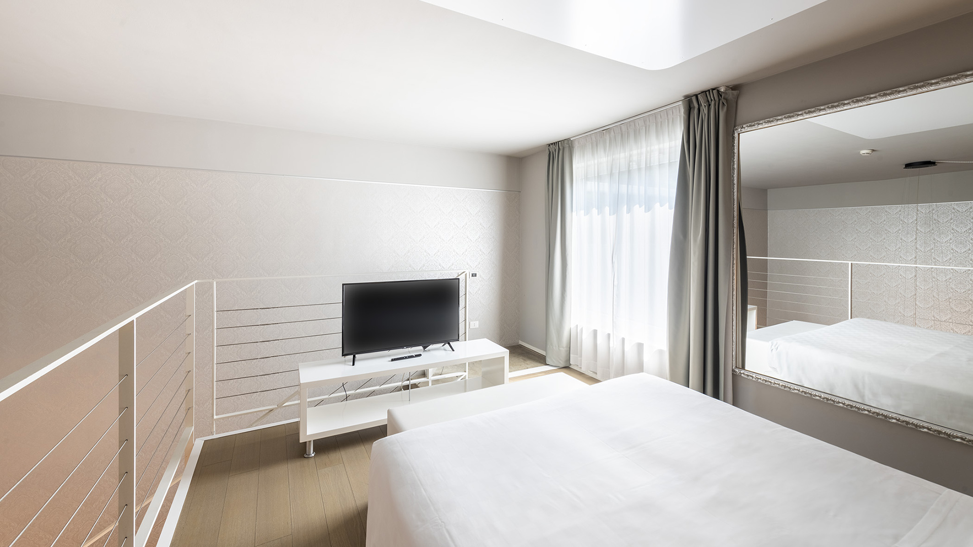 Junior Suite Deluxe su soppalco con letto matrimoniale, TV su mobile bianco, grande specchio a parete, pavimento in legno chiaro e ampia finestra con tende