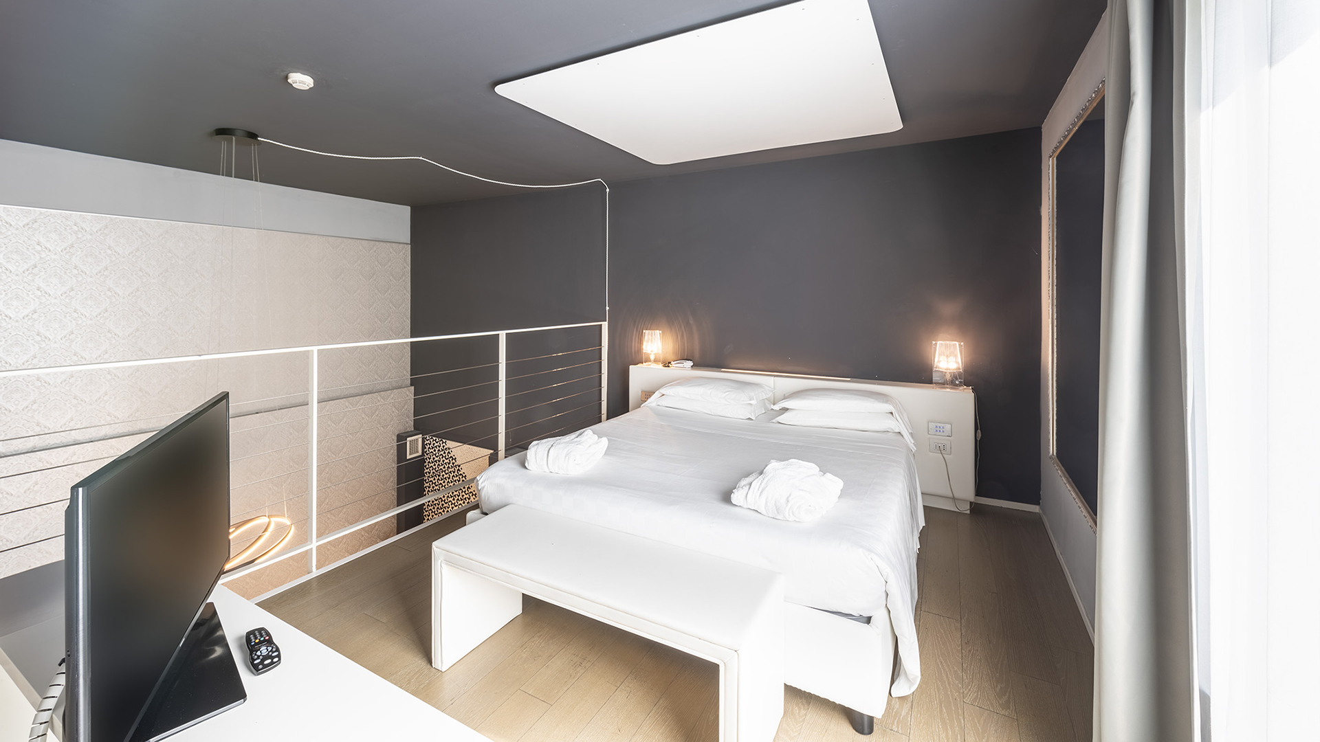 Elegante suite del MO.OM Hotel, luminosa con grande finestra, letto centrale, pareti scure e lampade di design