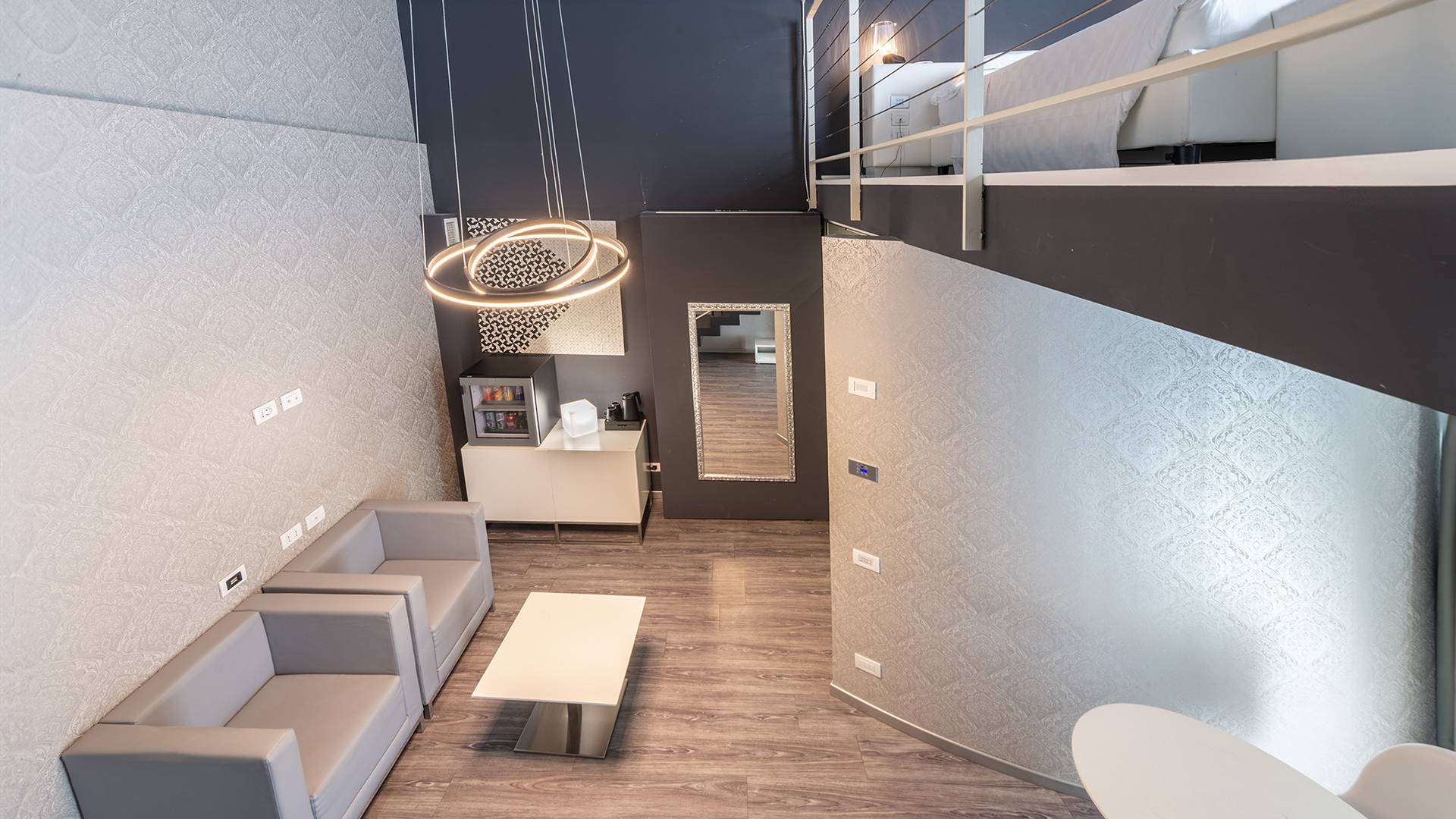 Lussuoso soggiorno di una suite a due livelli: pareti grigie, lampade circolari, pavimento in legno, due divani grigi e un tavolino bianco