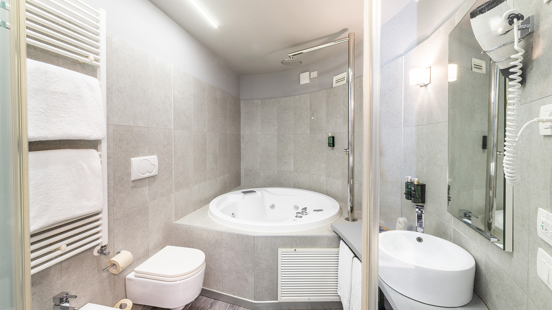 Suite con bagno dotato di vasca idromassaggio angolare, doccia a soffione, lavabo da appoggio, specchio illuminato e rivestimenti in gres effetto pietra