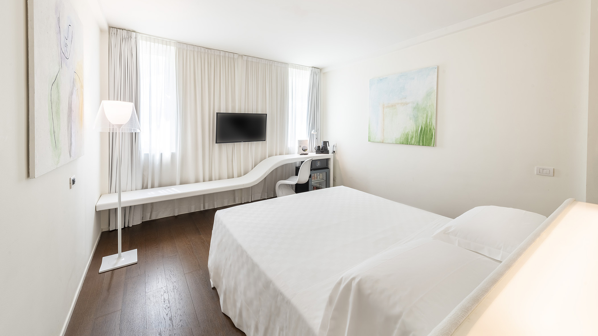 Camera Superior con letto matrimoniale, lunga panca bianca sotto la finestra con postazione scrivania integrata, TV a parete e ampie tende luminose